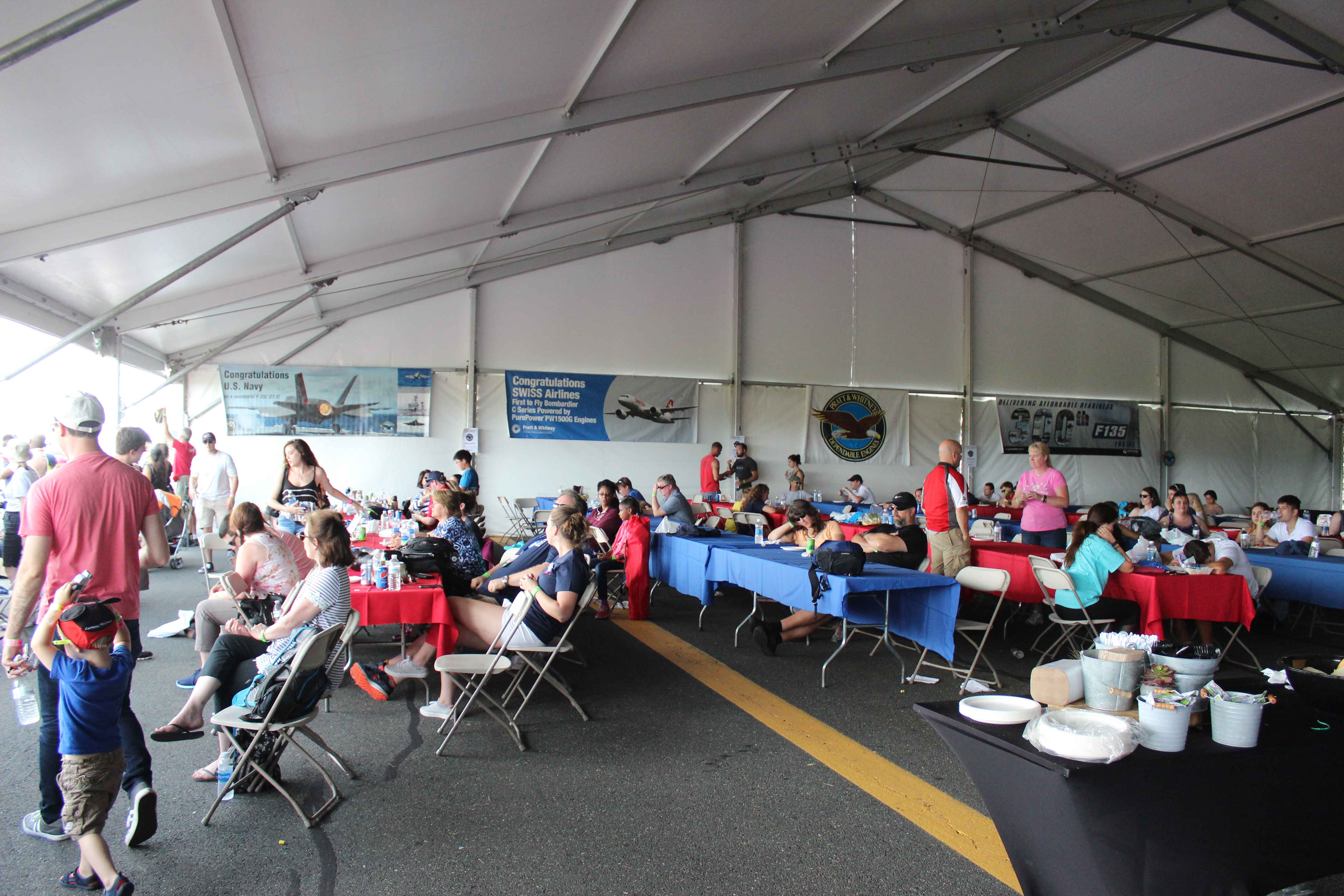 People inside a large white tent sitting at long tables watching an air show, there are posters for Pratt and Whitney hanging on the walls of the tent