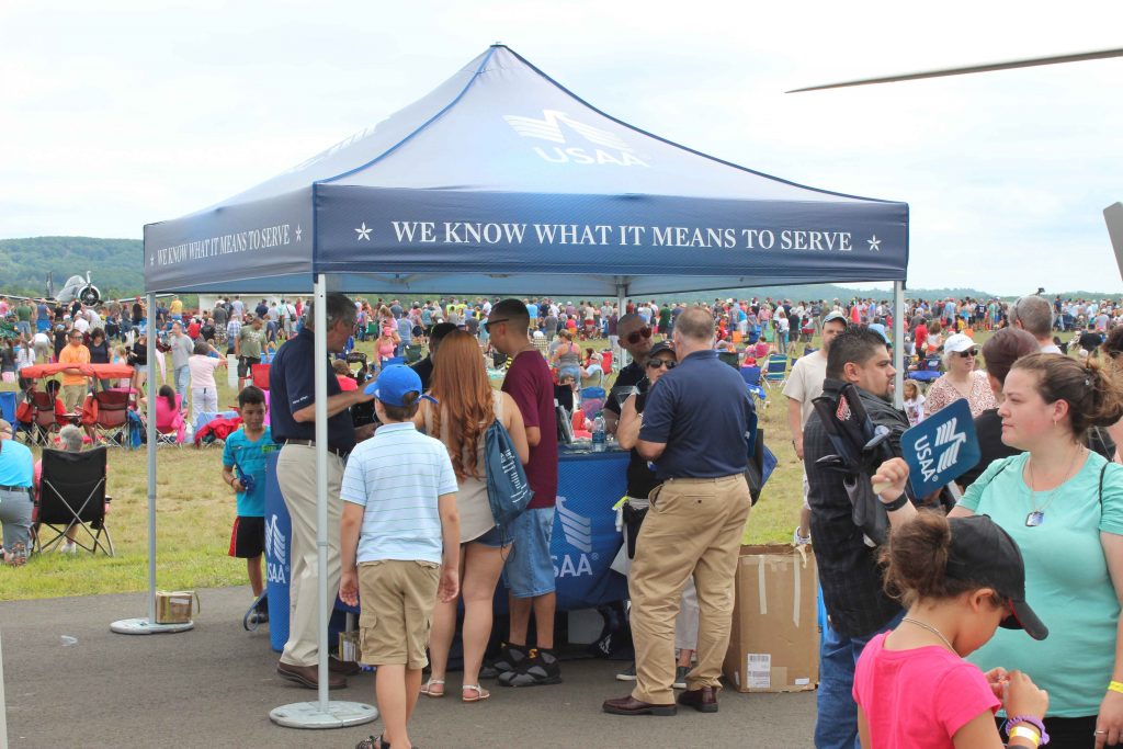 families visiting USAA exhibit under a branded navy blue pop up tent with a large crowd attending an air show in the background