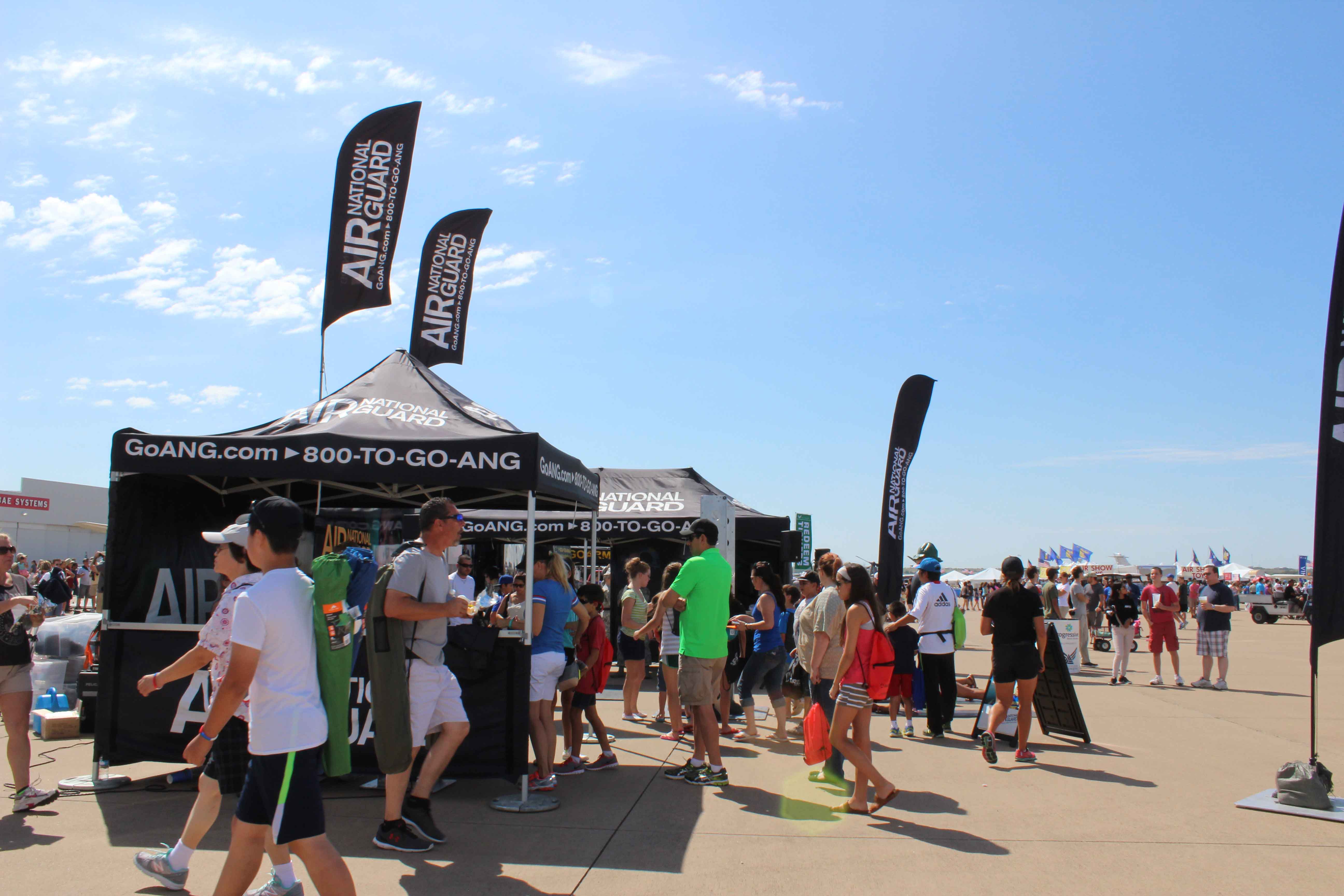 people around an Air National Guard exhibit with black branded tents at an air show on clear, blue sky day