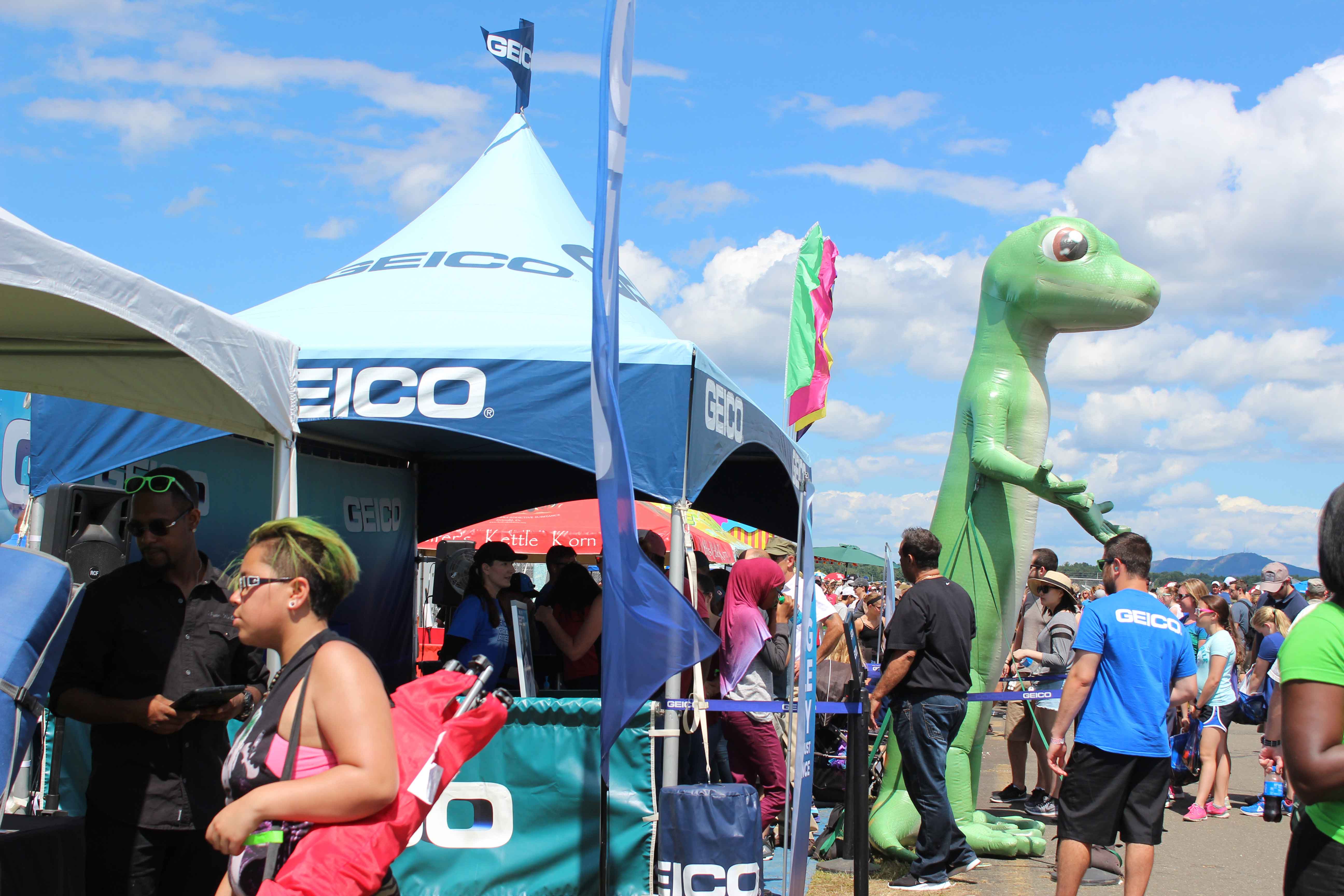 people exploring a GEICO exhibit that has a blue tent and a large blow up gecko in front of the tent at an air show on a beautiful day